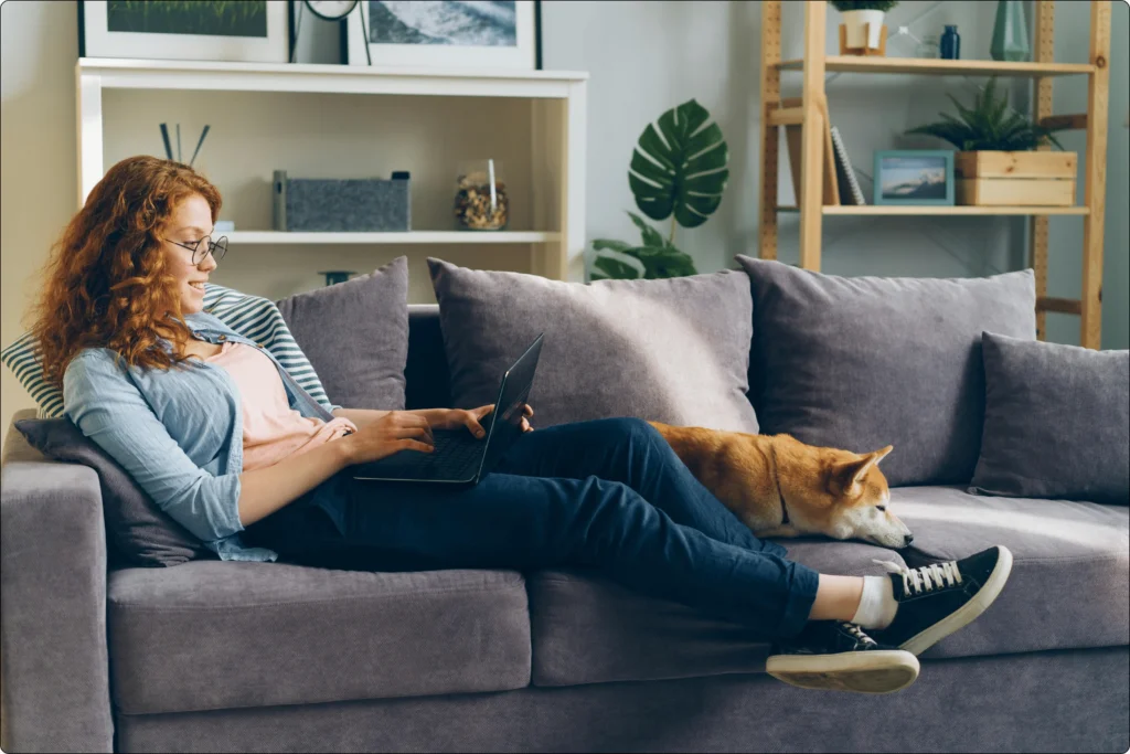 Comfortable and stylish: Woman wearing Judy Blue jeans and a light blue shirt, relaxing on a gray sofa with a laptop, accompanied by a dog, emphasizing the importance of comfort and versatility in Judy Blue denim.