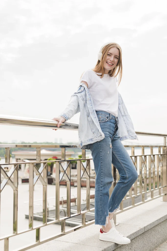 Young woman in branded Vervet Jeans and denim jacket smiling while leaning on a bridge railing, enjoying music outdoors with  her Superiority and Talent.

