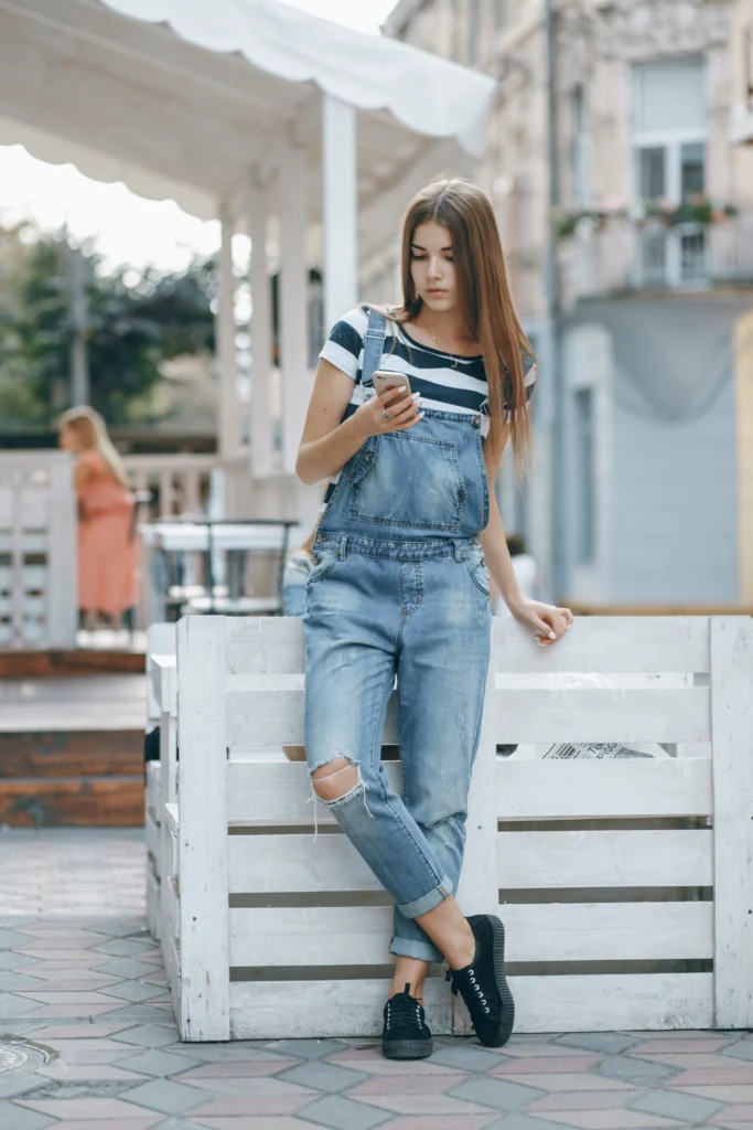 Young woman in distressed denim overalls and striped shirt leaning against a white wooden fence while checking her phone, showcasing the timeless appeal of bootcut jeans as a never-out-of-fashion style for casual urban looks.