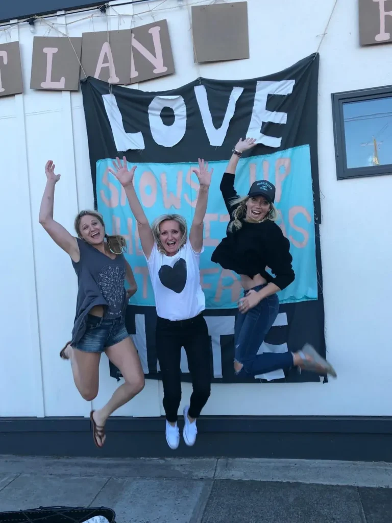 Three women jumping in front of a wall with a love-themed banner, wearing stylish flare jeans and casual tops. Celebrity-inspired casual look with trendy flare jeans.