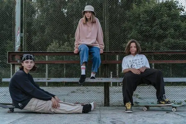  Three young people in casual streetwear, featuring hoodies, baggy jeans, and skateboards, relaxing at a skate park. The person in the center is sitting on a bench wearing a pink hoodie, blue jeans, and a bucket hat. The other two are seated on the ground, one in a black hoodie and beige pants, the other in a white t-shirt and black pants.