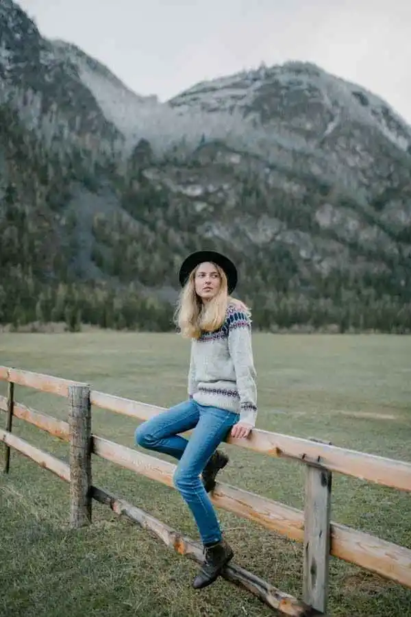 Woman wearing flare jeans, a cozy sweater, and a wide-brim hat, posing outdoors against a mountain backdrop. Showcasing stylish flare jeans women's flare jeans nature-inspired look