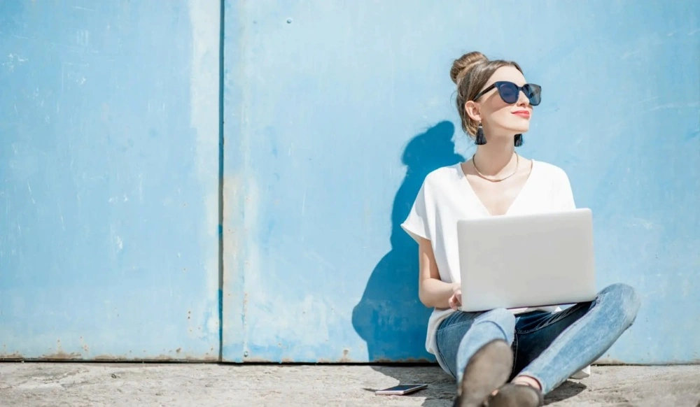 Woman in light blue jeans working on a laptop against a blue wall, representing women's pants size guide and finding the perfect fit.