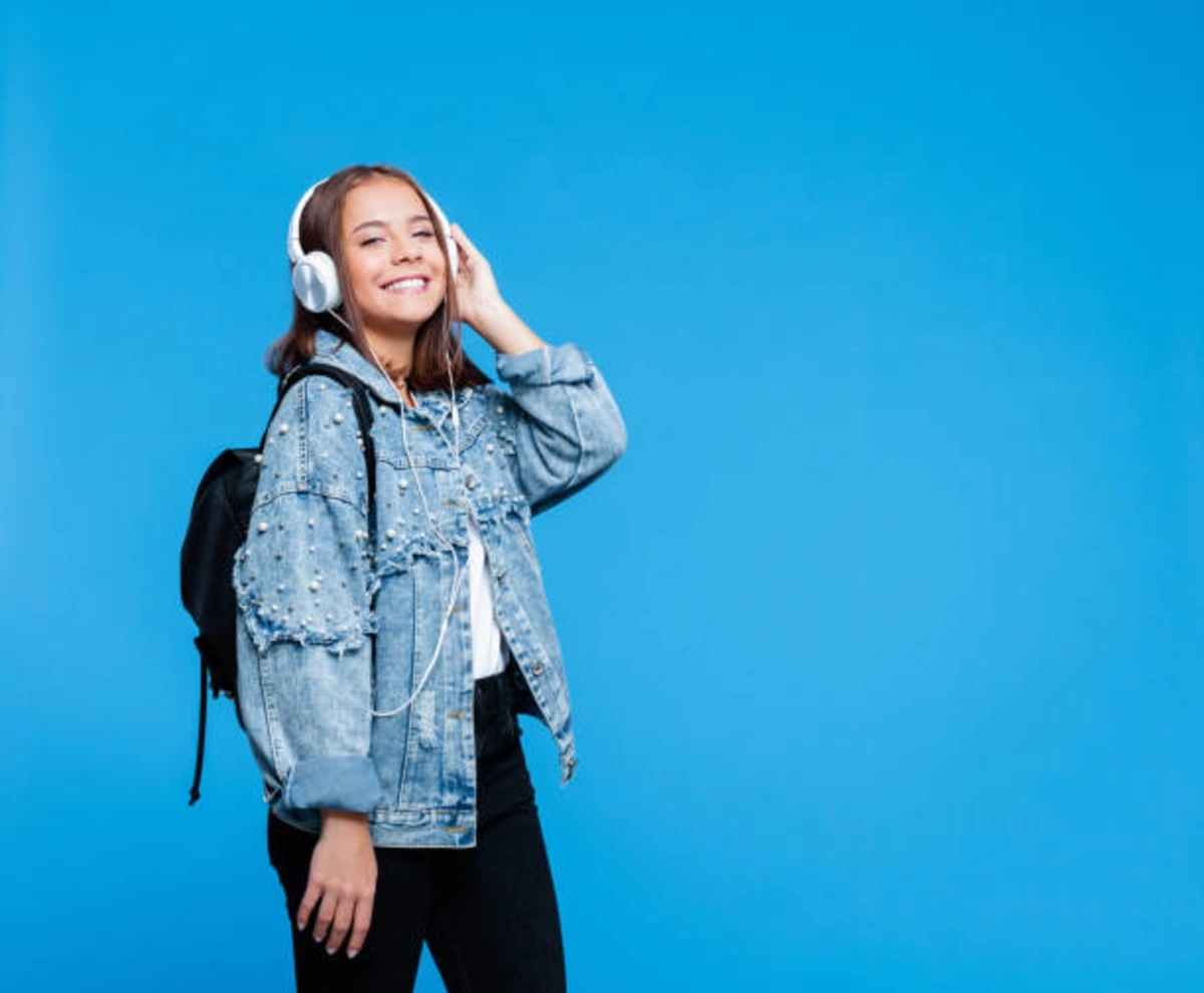 Young woman wearing a trendy denim jacket with pearl embellishments, black jeans, and headphones against a bright blue background.