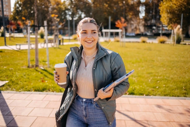 Young woman enjoying a sunny day outdoors, wearing a green sherpa-lined jacket with high-waisted Vervet jeans, holding a coffee cup and notebook, showcasing a cozy and stylish casual look perfect for fall