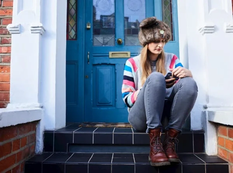 Young woman wearing mid-rise jeans paired with a colorful sweater and winter hat, sitting on steps outside a blue door.