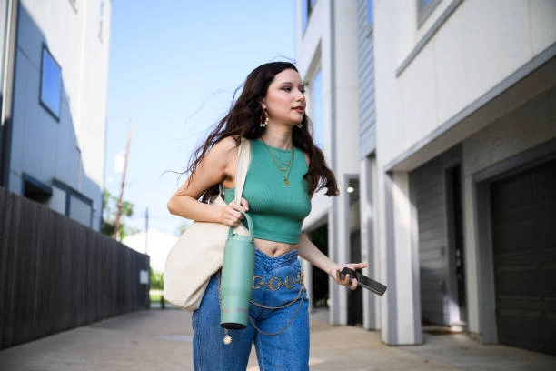 Fashionable young woman wearing medium wash bootcut jeans with a green crop top, accessorized with a belt chain, tote bag, and water bottle. This casual yet trendy outfit shows how to style medium wash bootcut jeans for a modern, everyday look for girls