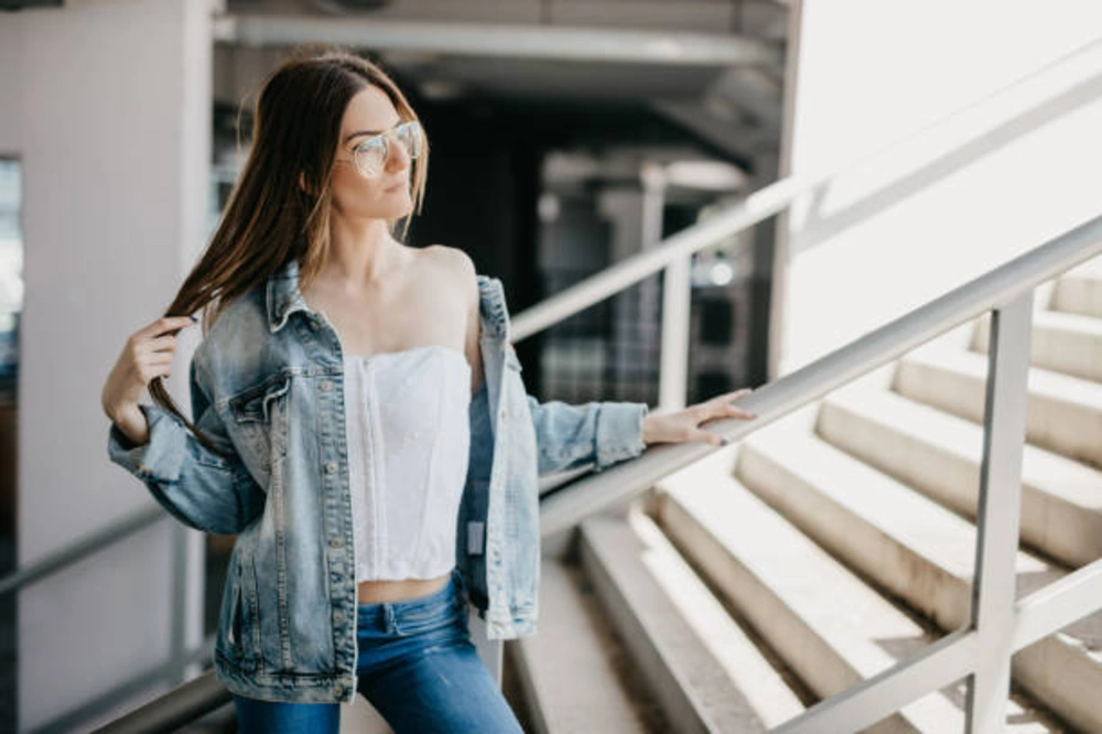 Fashionable young girl wearing women's blue jeans and denim jacket, styled with a trendy white top and sunglasses on a modern urban staircase.